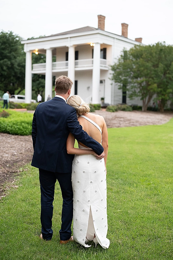 Couple portrait of bride and groom from behind, walking on a lawn toward a white columned estate, groom’s arm around bride in one-shoulder gown