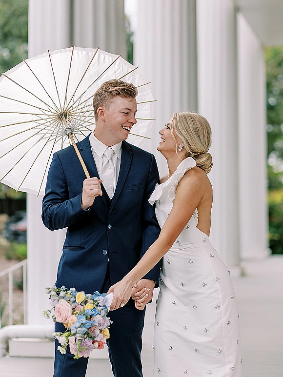 Couple portrait of bride and groom laughing under a white parasol, holding hands by white columns, her bouquet bright against his navy suit