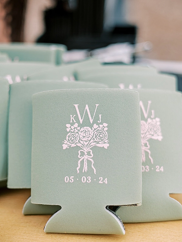 Wedding koozies and custom wedding koozies stacked on a wood tabletop, showing monogram print, floral illustration, and wedding date on foam sleeves