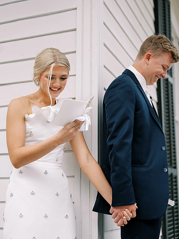 First look moment as bride in a strapless crystal gown reads a note card with ribbon, groom in suit waiting by white siding wall