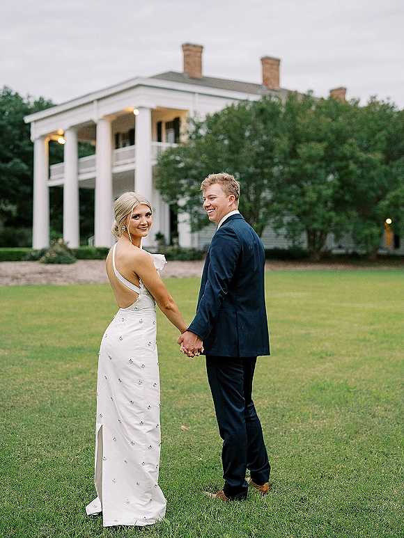 Couple portrait of bride and groom holding hands while walking away and looking back, bride in satin dress at manor house with white columns