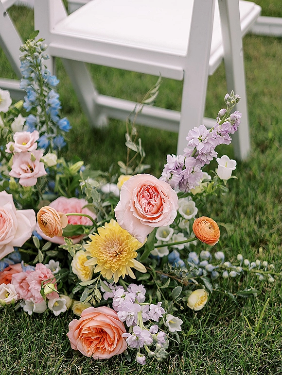 Aisle florals and wedding aisle flowers arranged low on a grass lawn with garden roses, ranunculus, delphinium, greenery by white chair