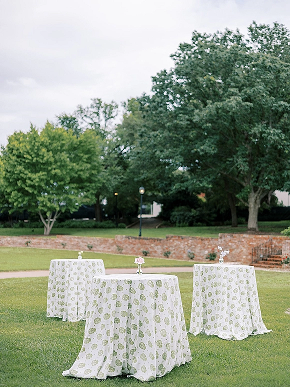 Cocktail hour setup with outdoor cocktail tables dressed in patterned linens and bud vases with single stem flowers on a grassy lawn by a brick wall