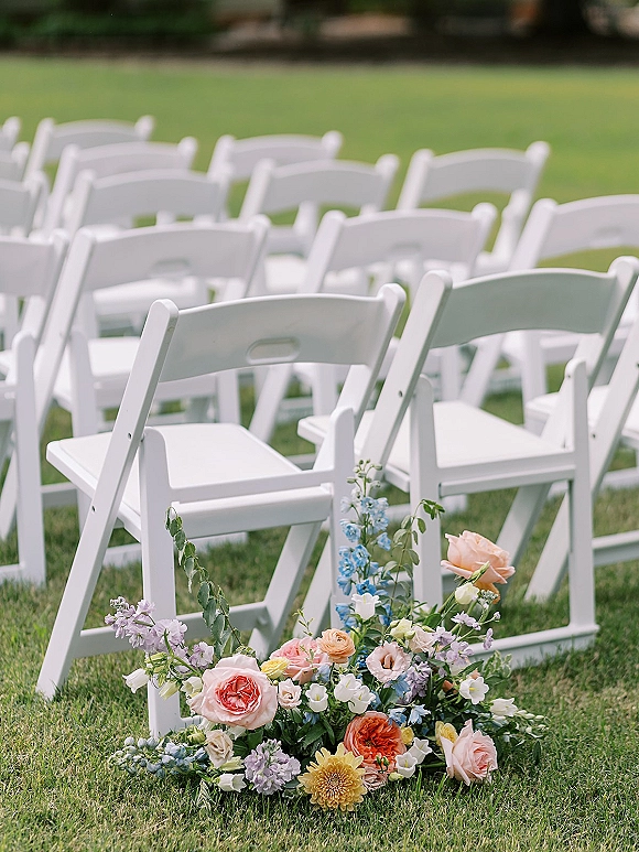 Ceremony seating with white folding chairs and low ground florals of roses, wildflowers, and greenery lining the aisle on a grass lawn