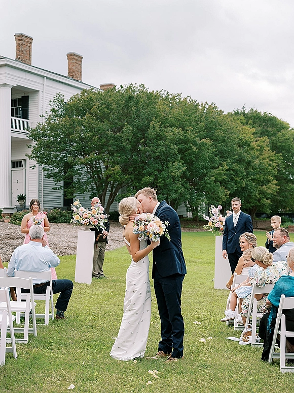 Wedding kiss as bride holds bouquet in a simple sheath dress and groom in navy suit, on a lawn aisle with petals, guests and white house backdrop