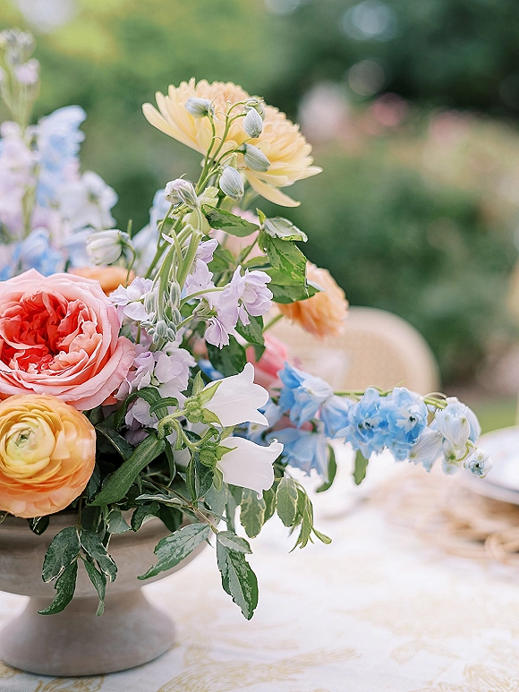 Wedding centerpiece of peach garden roses, yellow ranunculus and blue delphinium with greenery in a low stone compote on a linen table outdoors