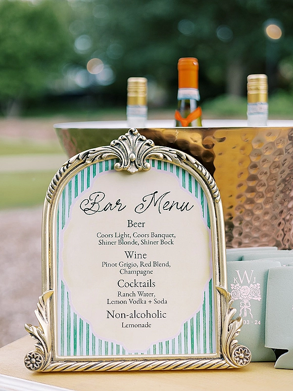 Wedding bar menu in an ornate gold frame with drink bottles, metal ice bucket, and cocktail napkins on a table with blurred greenery behind