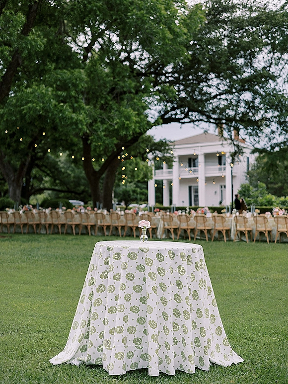 Outdoor reception setup with garden reception seating, round cocktail table and pink rose bud vase under string lights on an estate lawn