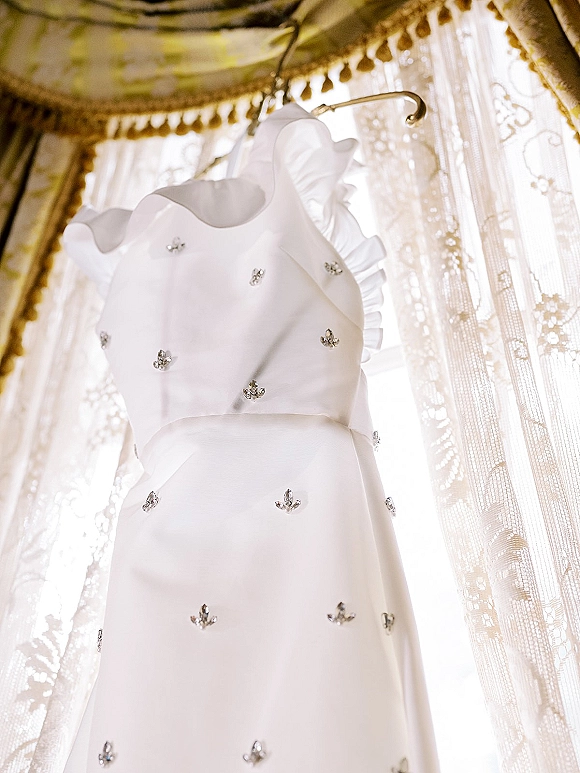 Wedding dress on hanger with rhinestone embellishments and ruffle trim, hanging by a sunlit window framed with sheer lace drapes
