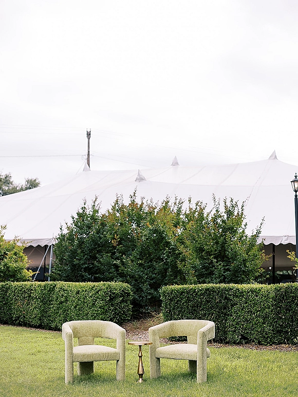Wedding lounge seating with two upholstered lounge chairs and a gold pedestal side table on a lawn beside trimmed hedges and a white tent