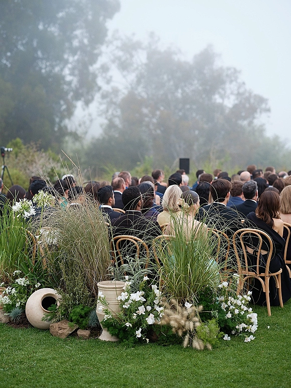 Outdoor wedding ceremony with wedding guest seating, wood chairs lining an aisle of white florals and grasses on a foggy lawn with trees
