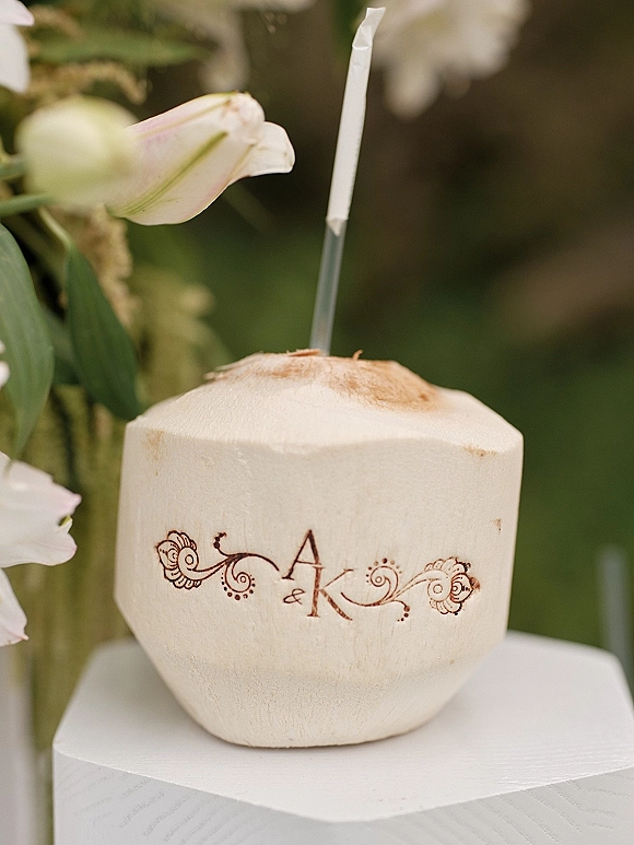 Wedding coconut drink in a fresh coconut with custom monogram print and paper straw, styled with floral accents on a table outdoors