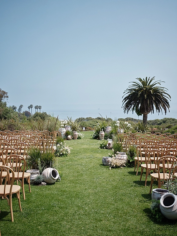 Ceremony aisle design with outdoor ceremony setup featuring wood chairs, white florals and pampas grass in urns on a lawn overlooking the ocean