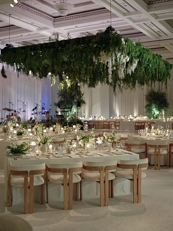Reception tablescape with long wedding tables in white linens, candlelit place settings, and hanging greenery under a draped ballroom ceiling