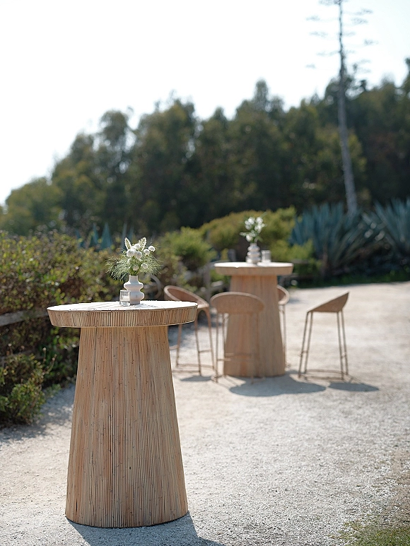 Cocktail table decor with white floral bud vases and glass votive candles on wood tables with bar stools on a gravel patio with agave plants