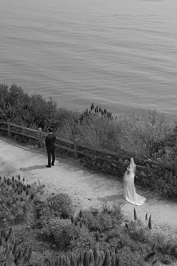 Wedding first look as bride approaches groom from behind in a long veil and dress train on a coastal bluff, ocean beyond a wooden fence
