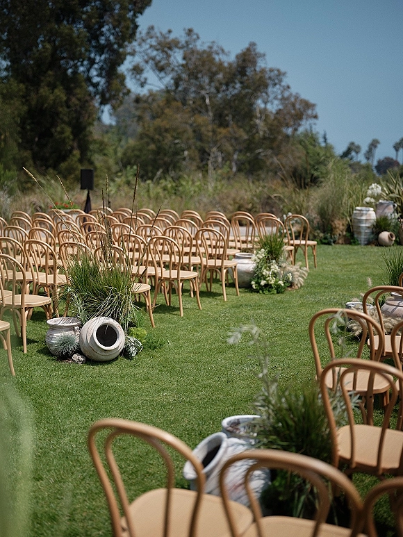 Ceremony setup with wood ceremony chairs lining an outdoor wedding ceremony seating aisle, pampas grass and urn florals on a grassy lawn