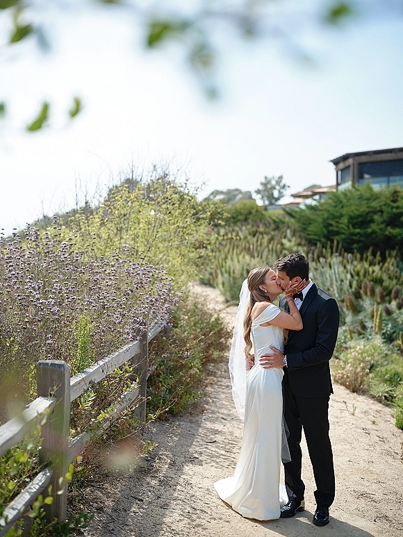 Wedding kiss portrait of the bride and groom kissing as she holds his face, her long veil flowing on a garden path by a wooden fence