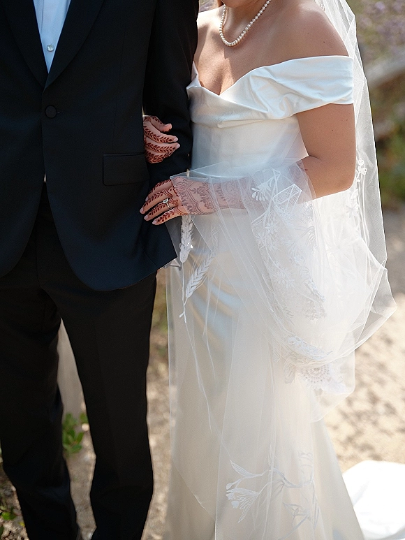 Couple portrait of bride holding groom’s arm, showing mehndi hands and engagement ring, with cathedral veil edge on a sunlit walkway