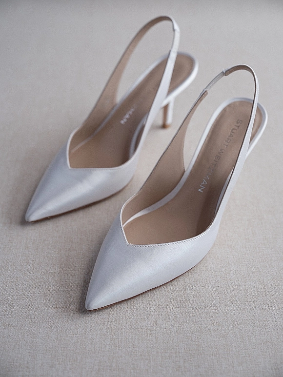 Wedding shoes in white bridal heels, satin slingback pointed-toe pumps arranged on a neutral fabric surface in a flat lay