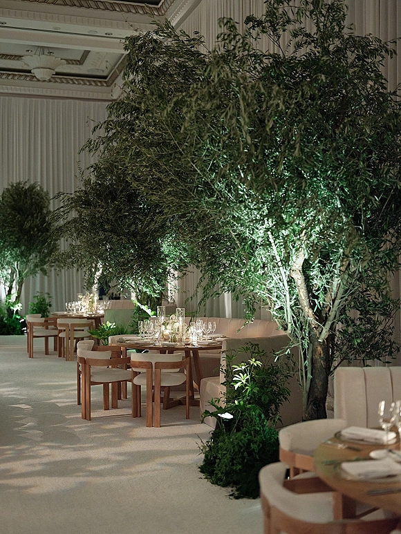 Reception tablescape with round wooden tables, candlelit glassware and linen napkins, set among indoor trees in a coffered ballroom