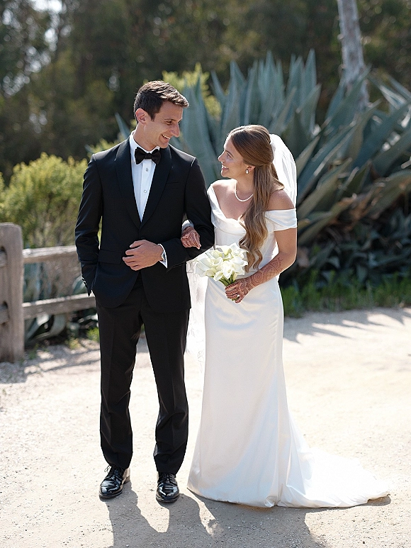 Couple portrait of bride and groom walking arm in arm, she holds white calla lily bouquet, in a garden with agave and trees