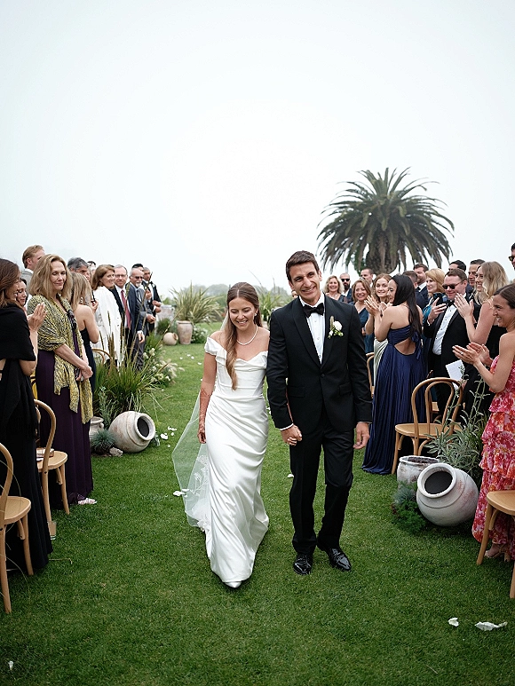 Wedding recessional as bride and groom walking aisle hand in hand, cathedral veil flowing, guests cheering on a garden lawn with palm tree backdrop