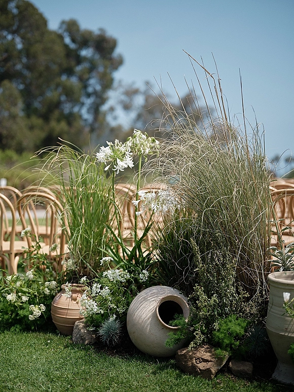 Ceremony aisle decor with terracotta pots of ornamental grasses, white flowers and greenery lining a lawn aisle beside wooden chairs under blue sky