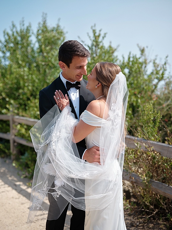 Couple portrait of bride and groom embrace, her wedding veil blowing with lace edge as they hold close by trees and a wooden fence