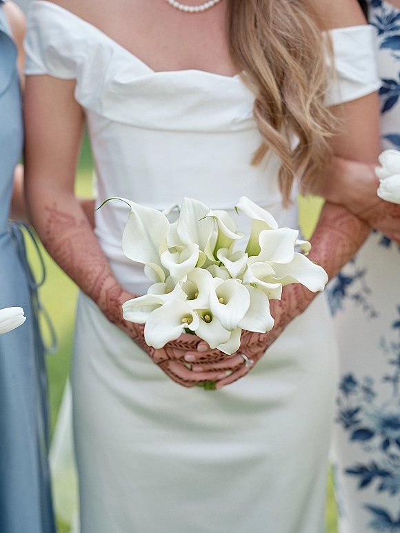 Bridal bouquet of white calla lilies cradled in a bride’s hennaed hands with a wedding ring, greenery and bridesmaids in blue behind