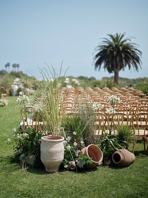 Ceremony aisle decor with terracotta urn floral arrangements and pampas grass beside wood chairs on a lawn with a palm tree backdrop