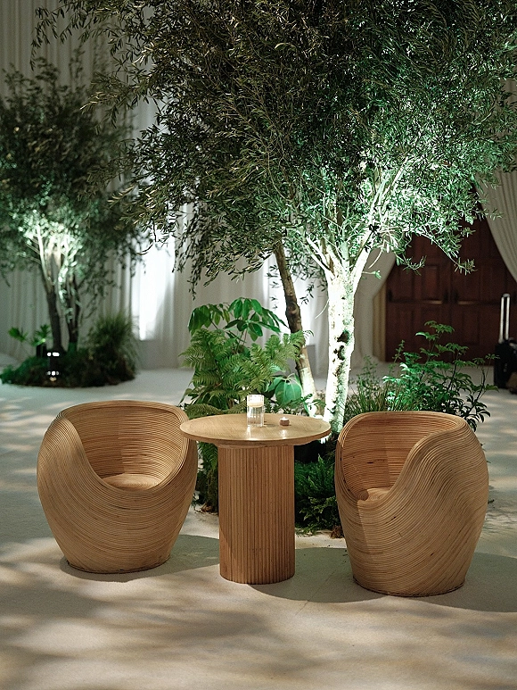 Wedding lounge seating with wood lounge chairs around a round pedestal table, candlelight, potted trees, ferns, and white drapery indoors