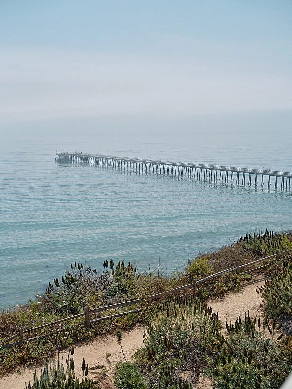 Ocean pier stretching over water with a wooden fence and dirt path along coastal shrubs, facing a calm horizon by cliffside coast