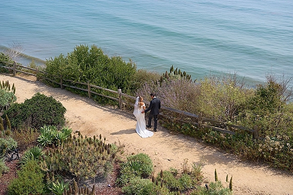 Couple portrait of bride in wedding dress and veil with groom in suit holding hands on a coastal bluff path by ocean and fence