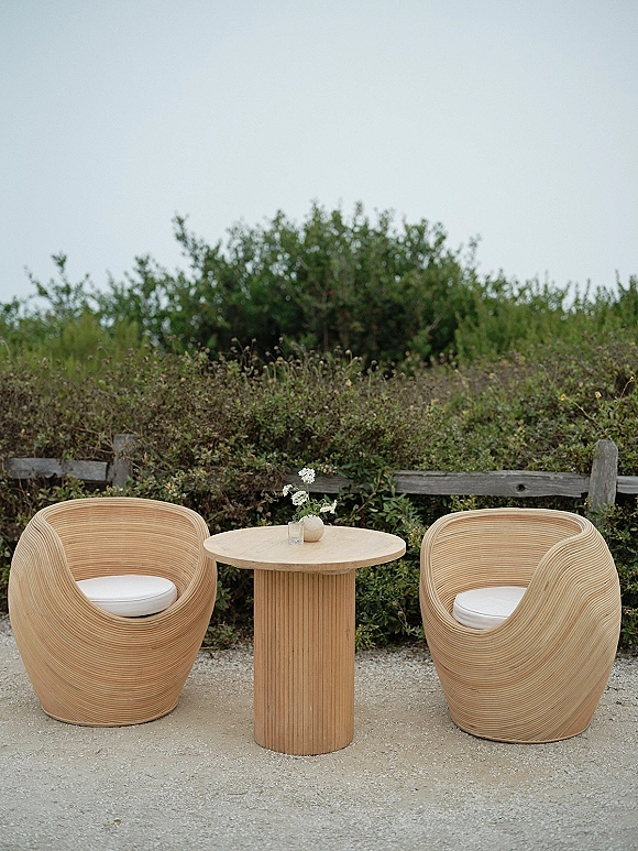Outdoor lounge seating with woven chairs around a round wood table, accented by a small vase of white flowers on gravel by a wood fence
