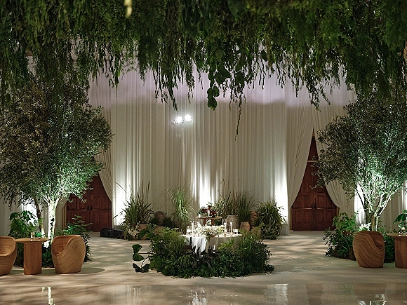 Ceremony backdrop with white draped backdrop, lush greenery and potted plants, candlelit lanterns and lounge seating in an uplighted hall