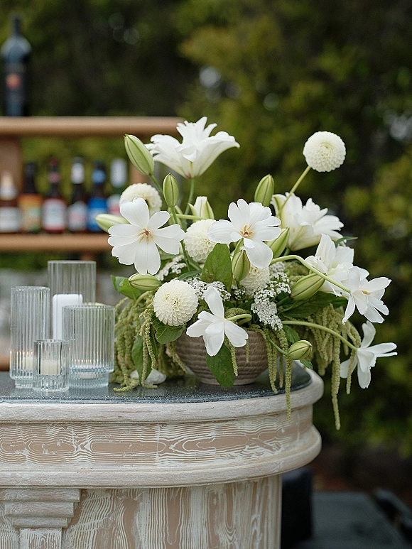 Wedding bar decor on a wooden outdoor wedding bar with white floral centerpiece, glass candle holders and liquor bottles amid green trees