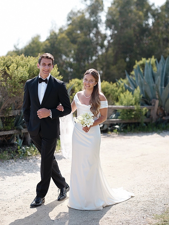 Couple portrait of bride holding calla lilies beside groom in black tuxedo, her veil and pearls glowing along a garden path