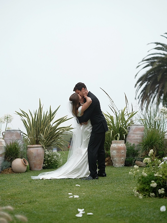 Wedding kiss as the bride and groom kissing on a garden lawn, veil and dress train flowing beside terracotta urns and petal aisle