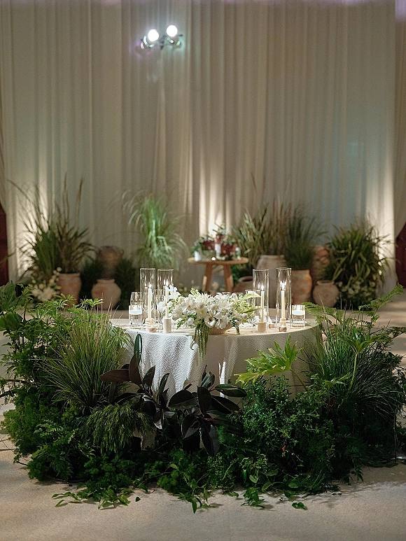 Wedding sweetheart table with sweetheart table decor, white linens, low white florals, greenery garland, and pillar candles before draped backdrop