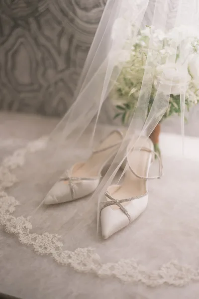 Bridal shoes with white wedding heels and rhinestone straps styled beside a tulle veil and rose bouquet on a neutral floor in soft light