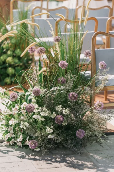Ceremony aisle florals with aisle ground florals of white blooms, greenery, and purple allium lining chairs on a sunlit stone patio aisle