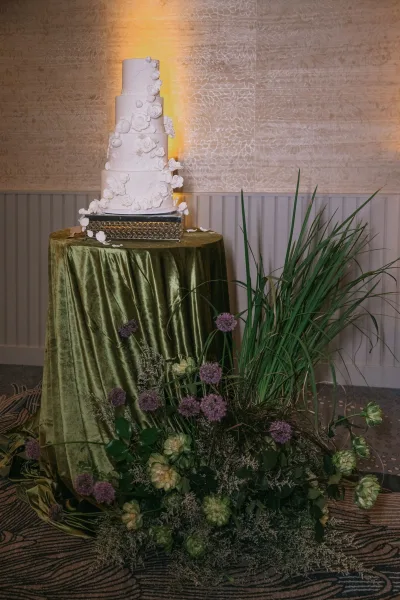 Wedding cake four tier white design with cascading sugar flowers on a stand, styled with purple florals and greenery against uplit wall
