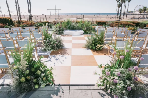 Ceremony setup with beach wedding ceremony seating, gold chairs and checkered aisle runner leading to a round platform by the oceanfront shore