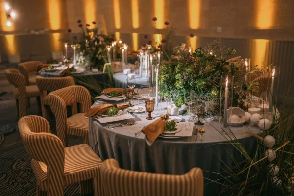 Reception tablescape with round wedding table setup featuring greenery centerpieces, tall taper candles, amber goblets, and gray linens in a ballroom with uplighting
