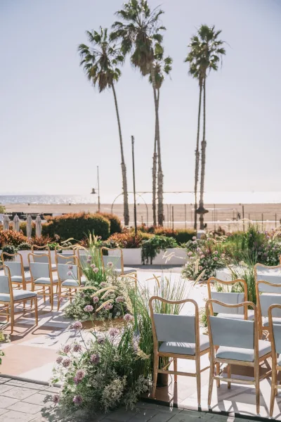 Ceremony setup with outdoor wedding ceremony chairs lining a flower-filled aisle, framed by palm trees and oceanfront patio views under blue sky