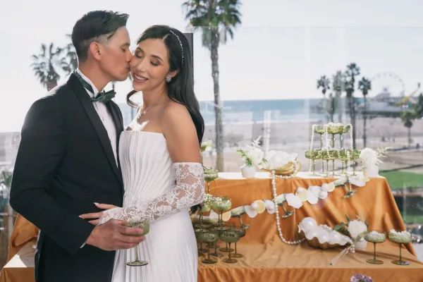 Wedding couple portrait of bride and groom toast as he kisses her cheek beside a champagne coupe tower on an oceanfront terrace with palms