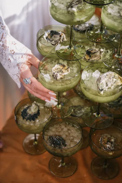 Oyster champagne tower with green coupe glasses and oysters on ice, bride’s lace-sleeved hand showing an emerald cut ring on a wood table