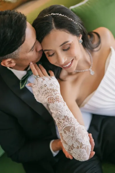 Wedding couple portrait with groom kissing bride’s cheek as she closes her eyes, lace gloves and ring visible on green upholstered seating