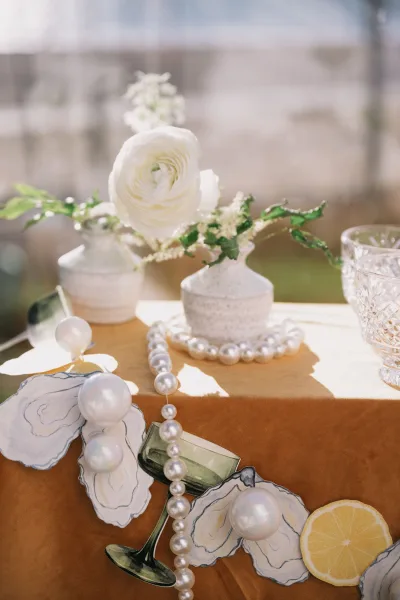 Wedding tablescape with oyster shell place cards, bud vases of white flowers, pearl necklace, crystal goblet, and lemon slice in soft daylight